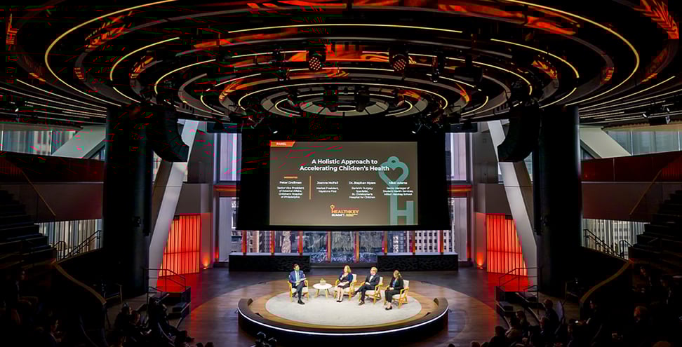 Panel discussion at the HealthKey Summit with four speakers on stage beneath a large screen titled “A Holistic Approach to Accelerating Children’s Health” in a modern auditorium with dramatic circular lighting.