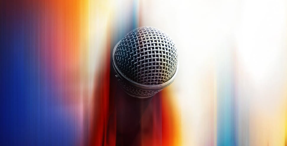 A close-up of a microphone against a blurred, colorful background with vertical streaks of blue, red, orange, and white, creating an abstract and dynamic visual effect.